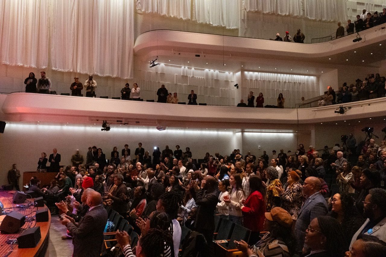Audience in a modern auditorium giving a standing ovation, with two balconies filled with standing spectators, dim lighting, and a stage visible on the left.